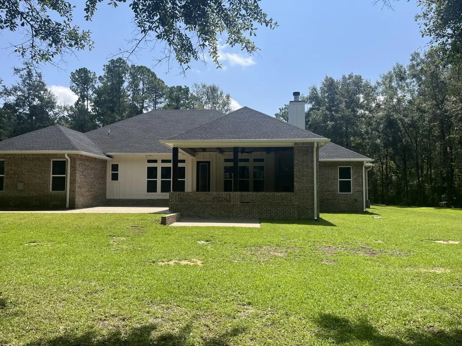 Rear of the brick home with a covered, screened back porch and a large mowed lawn bordered by pines